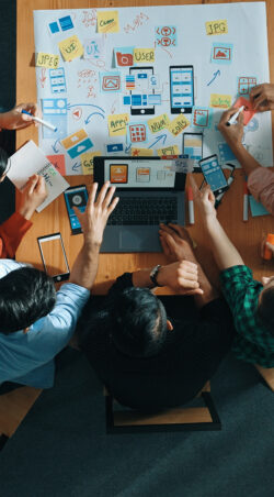 Top view of skilled manager using laptop working on Ux Ui design. Convocation. Top view of project manager using laptop working on Ux Ui design for mobile phone interface while marketing team designing software program by using brainstorming mind map. Top view. Convocation.